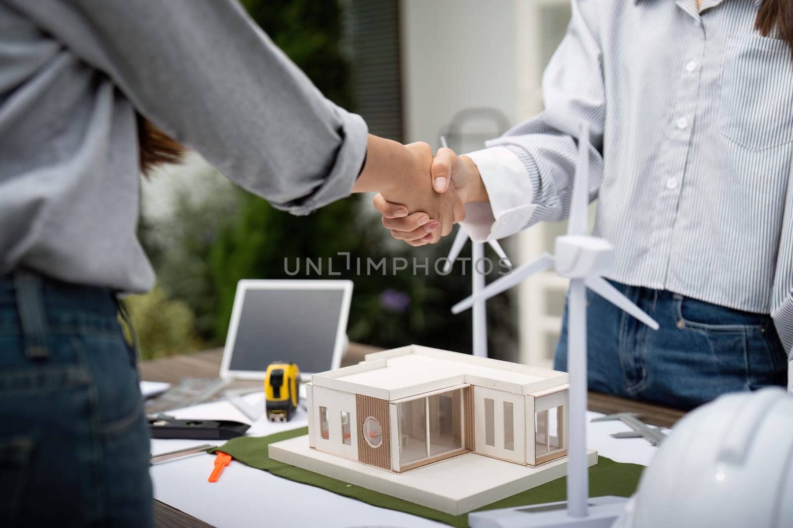Two sustainability engineers shaking hands as a sign of agreement on their project, with architectural models and blueprints on the table outdoors.