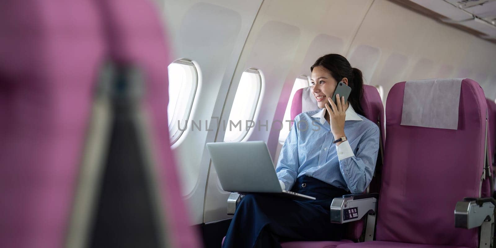 A professional Asian woman using a laptop and talking on the phone while seated on an airplane, embodying productivity and comfort during business travel.