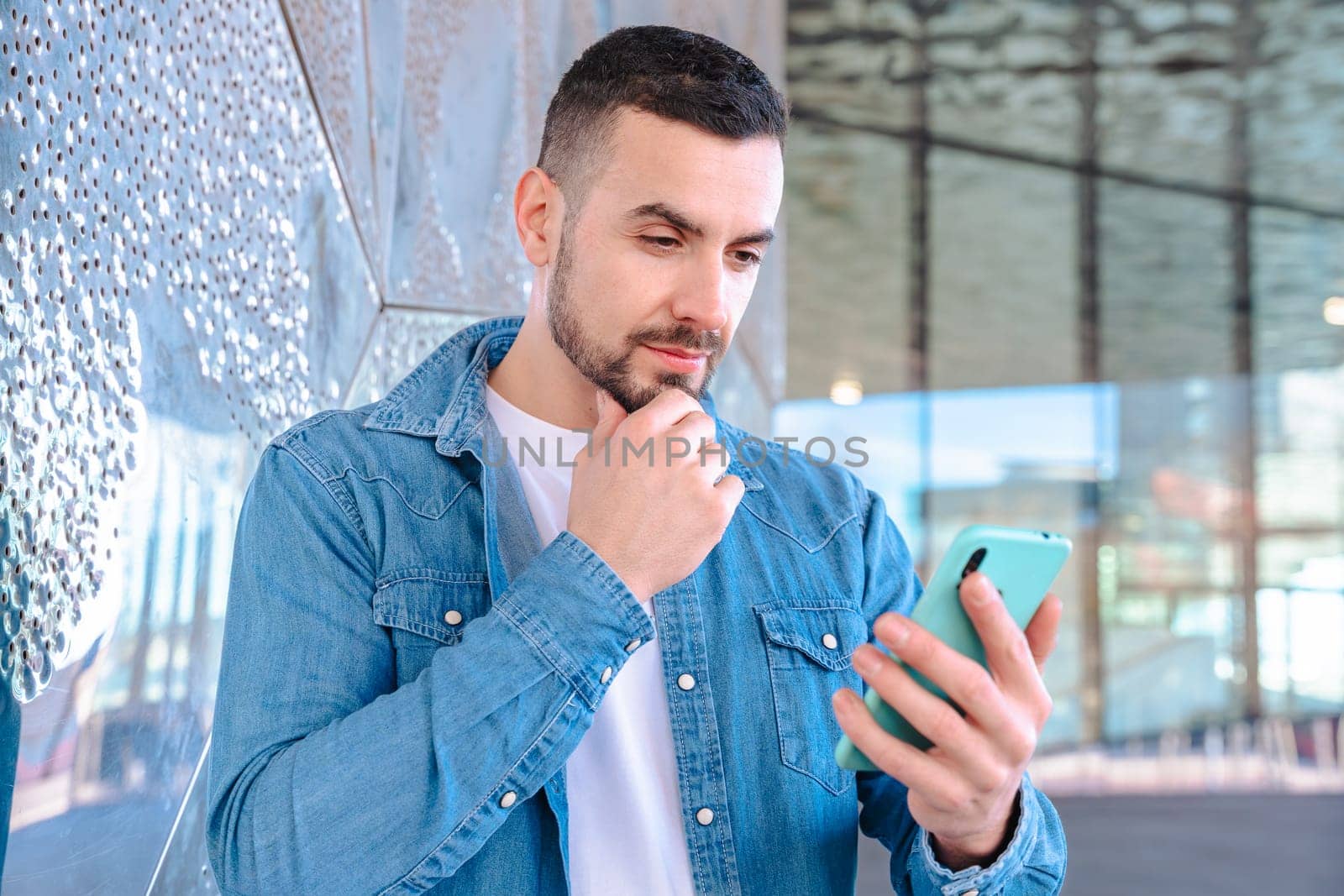 young man wearing a denim shirt, standing outdoors near a reflective glass wall, thoughtfully looking at his smartphone in natural daylight, symbolizing communication and modern life.