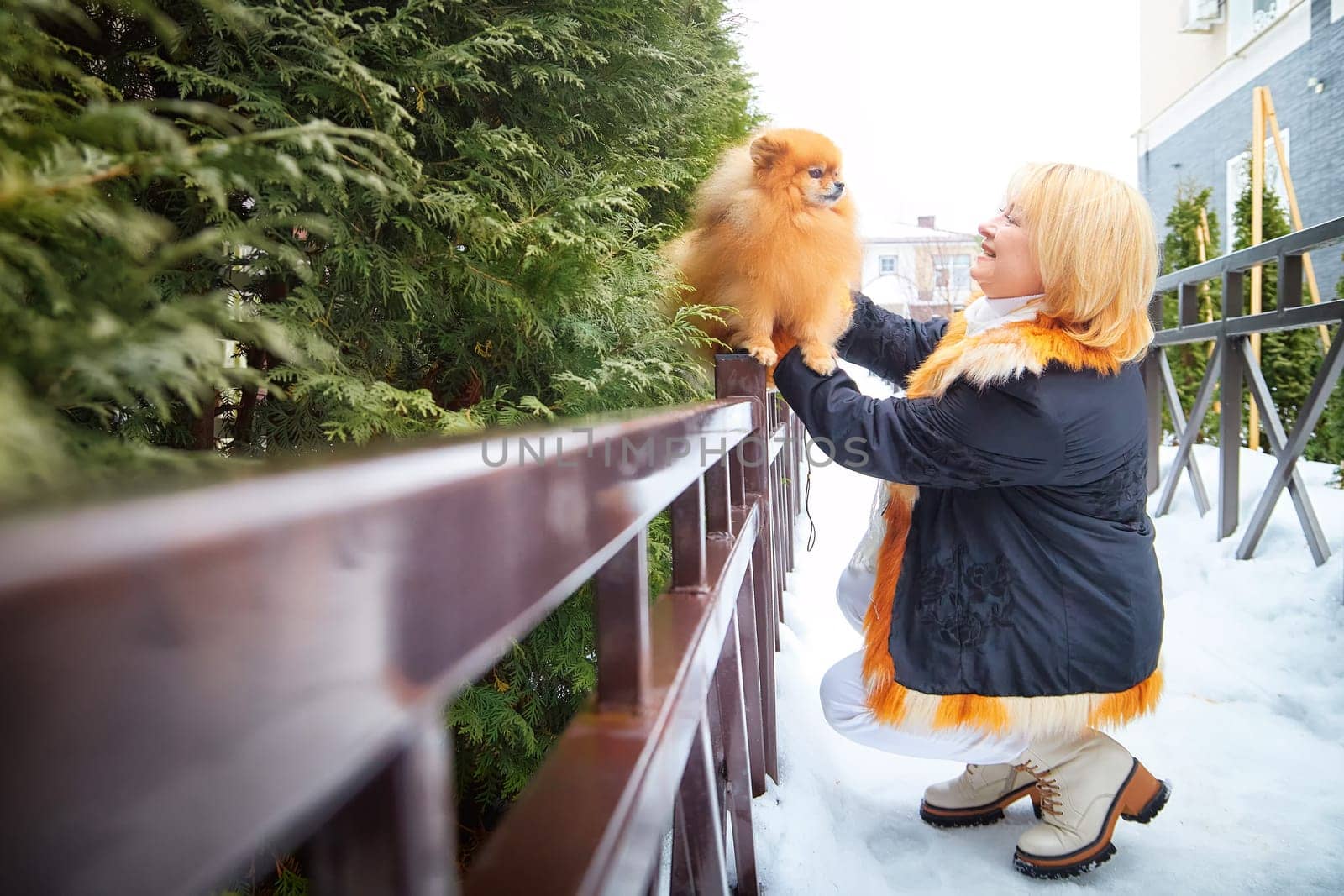 A middle-aged woman lovingly lifts her fluffy Pomeranian while enjoying a snowy winter day outdoors.