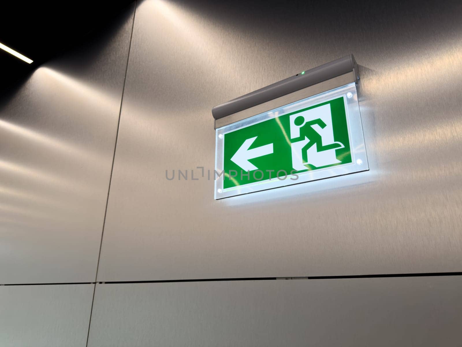 Glowing green emergency exit sign with arrow and running person symbol on brushed metal wall. Evacuation indicator in modern public building. Concept of alert, escape route and safety compliance