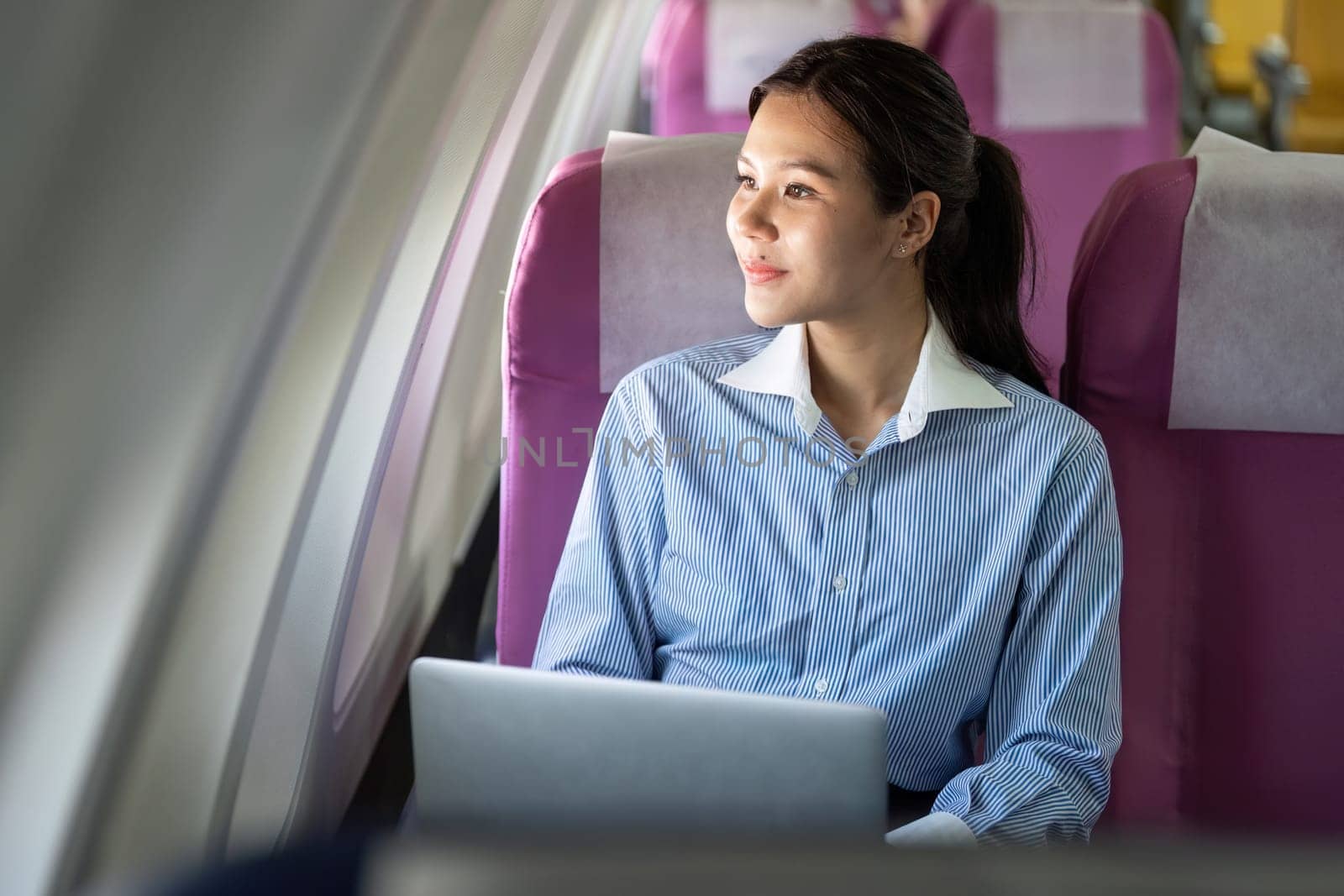 A young professional woman smiles while using her laptop on an airplane, showcasing the convenience of business travel.