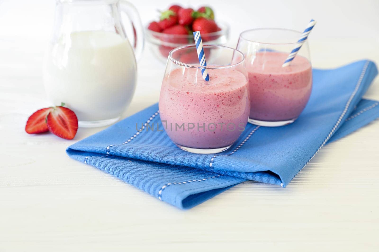 Two colorful strawberry smoothies in clear glasses are placed next to a pitcher of milk and a bowl of fresh strawberries on a blue napkin.
