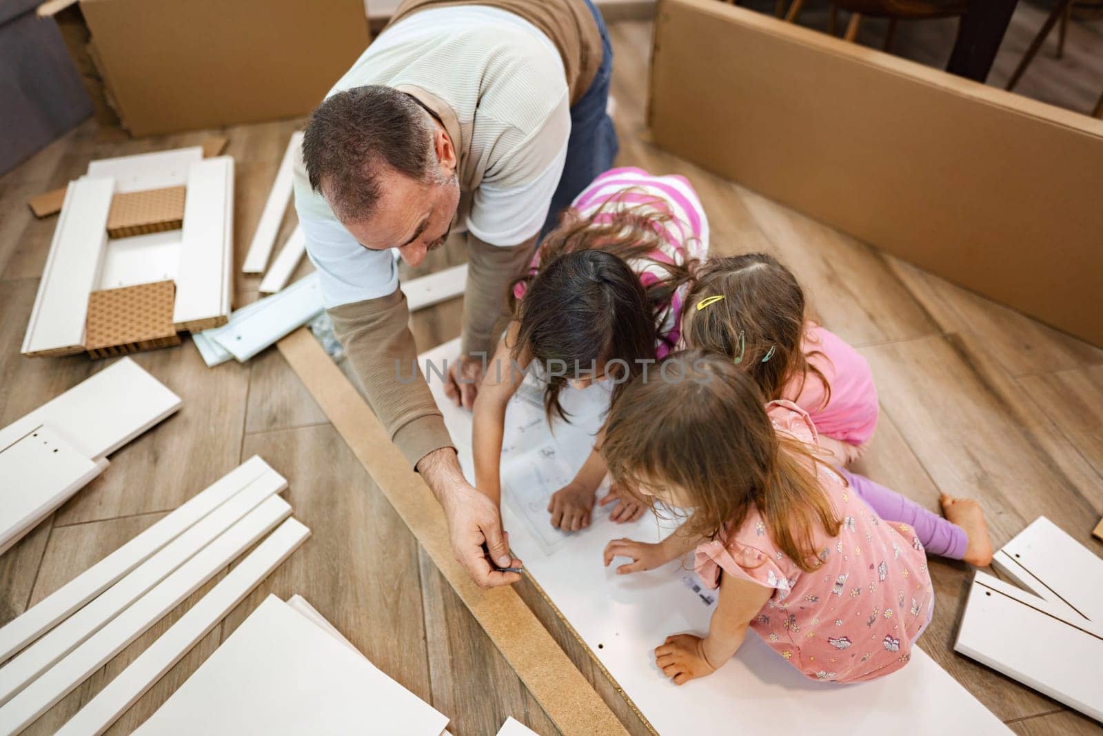 A delighted father guides his three young daughters as they assemble a furniture piece in a warm, inviting room filled with natural light and laughter. Each child contributes to the activity.