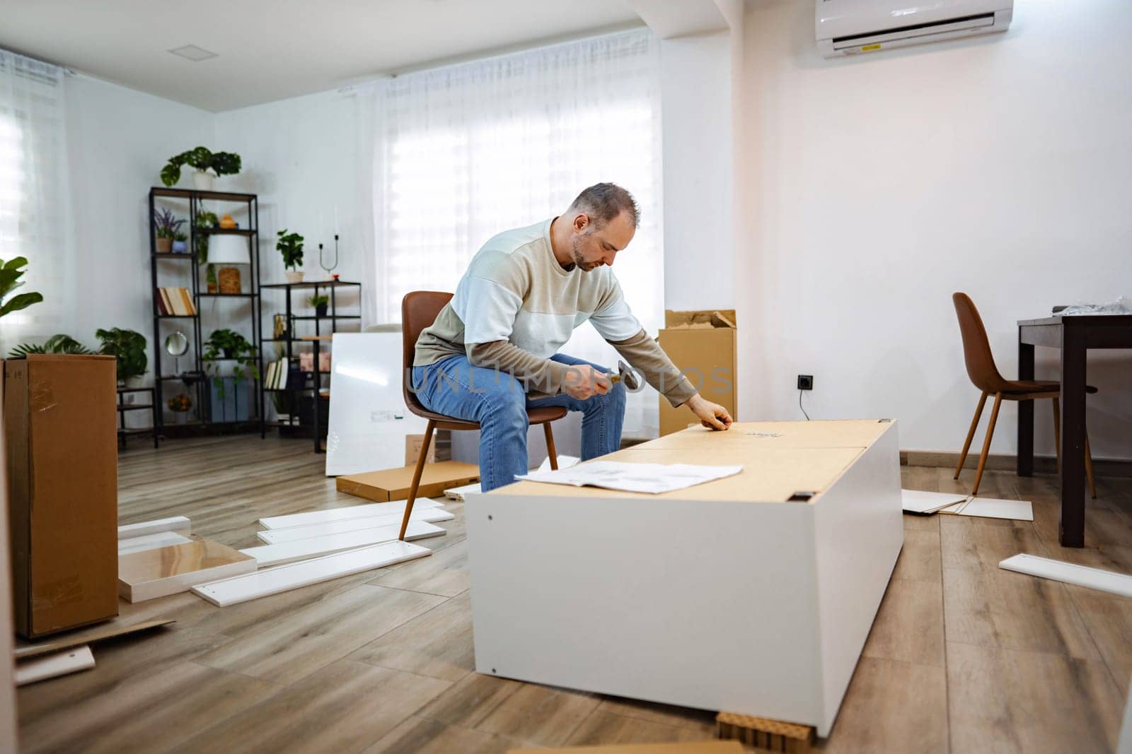 In a cozy, well-lit room, a focused man is piecing together furniture. Surrounded by packaging materials, he meticulously examines each component, creating a welcoming atmosphere.