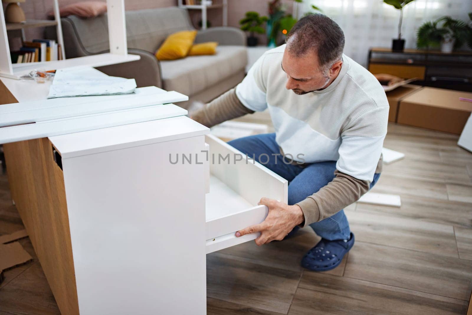 A man focuses intently as he fits a drawer into a newly built piece of furniture in his stylish living room. Surrounding boxes and tools hint at a productive day of assembling.