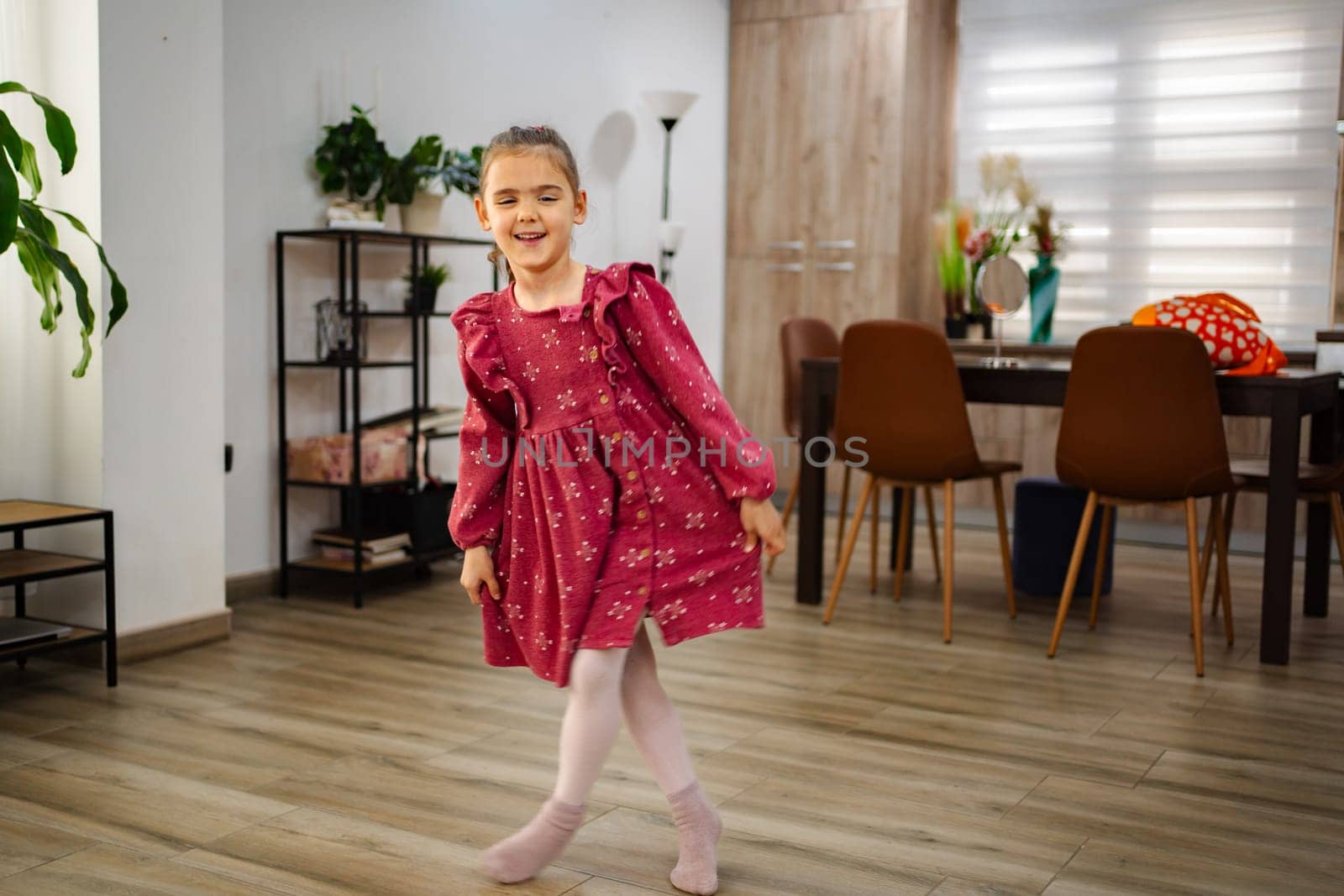 A cheerful young girl twirls gracefully in a cozy living room, wearing a lovely pink dress. Sunlight floods the space, highlighting a warm atmosphere filled with plants and furniture.