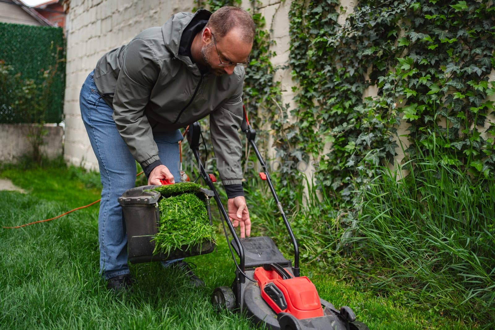 Gardener focuses on maintaining his vibrant lawn, emptying freshly cut grass clippings from a mower into a bucket. The surroundings are lush, highlighting a peaceful evening atmosphere.