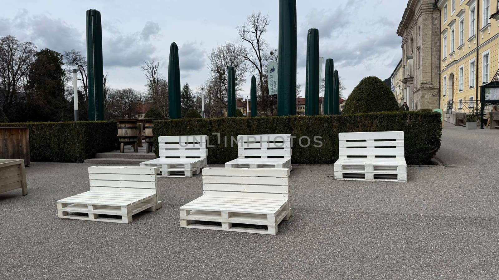 Tegernsee, Germany - March 30, 2025: Outdoor seating area features white pallet furniture in a park for relaxation near lake Tegernsee and church Pfarrkirche St. Quirinus