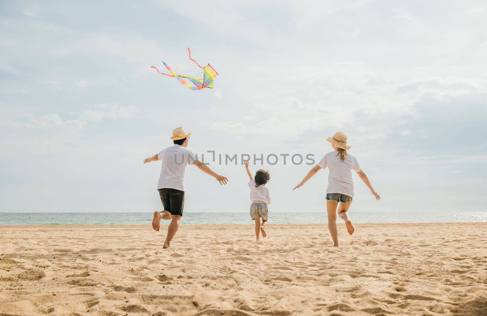 Asian family is flying a kite high in the sky while running on the beach. Happy parents mother and father with their child playing with kite, family is enjoying their time together, summer day
