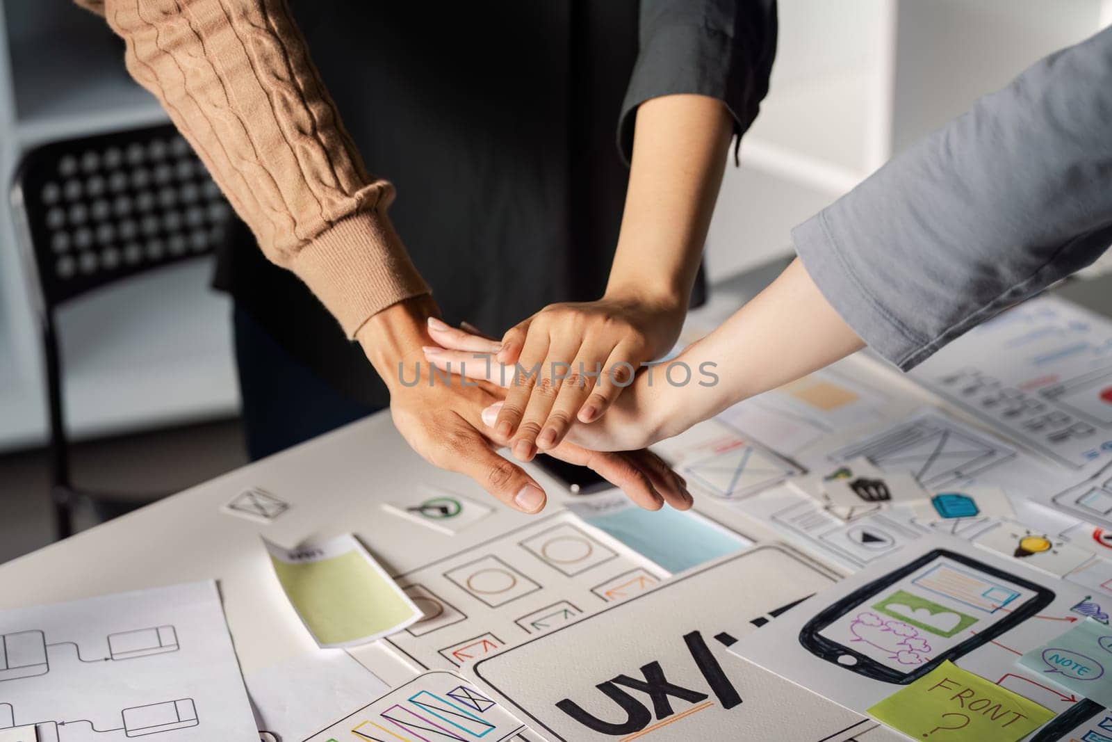Team members joining hands in solidarity while engaged in a UX UI design collaboration, surrounded by sketches and visual materials on the table.