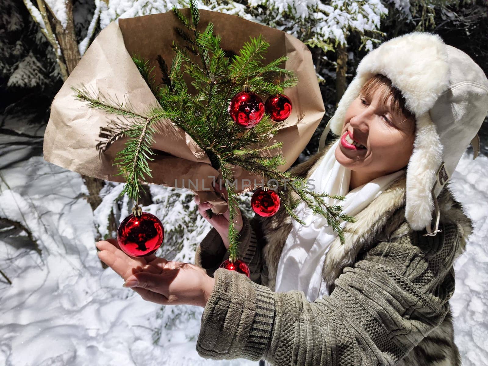 Middle-aged woman dressed warmly is having fun during a night walk in the snow-covered forest, playfully showing festive decorations.