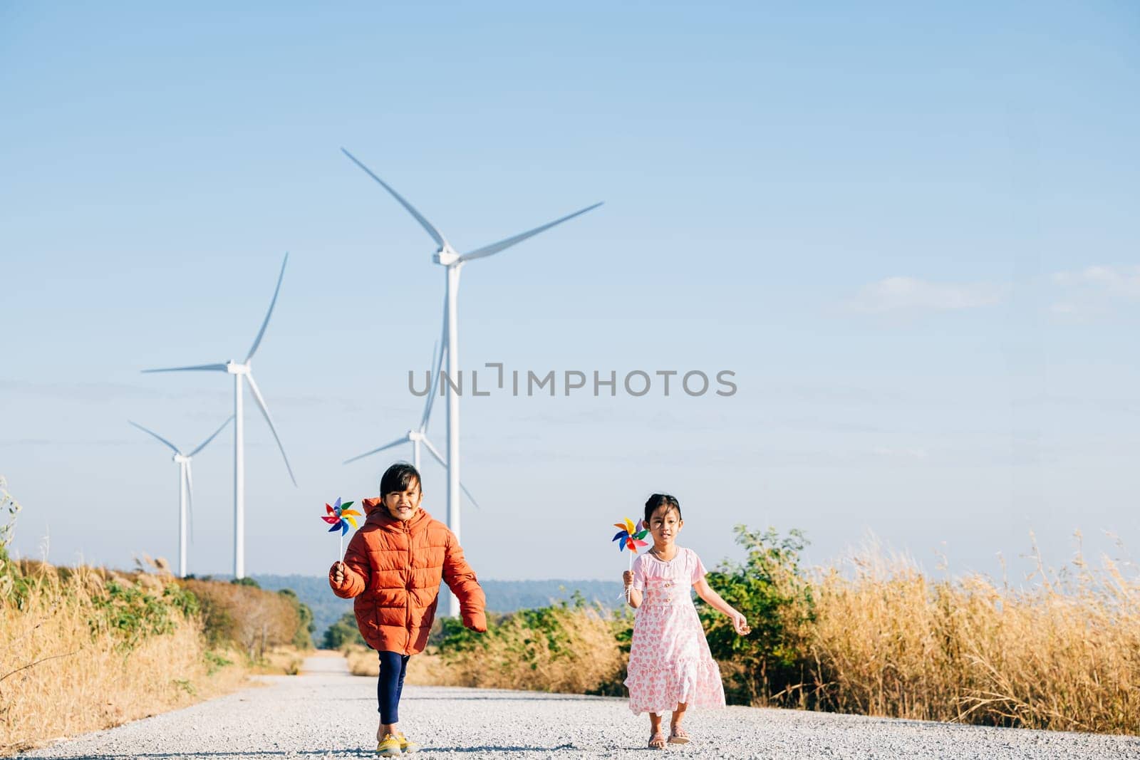 A girl and her sister holding pinwheels run by windmills. Family bonding near turbines signifies clean energy. Children joy windmill circle against clear sky reflects sustainable cheerful future.