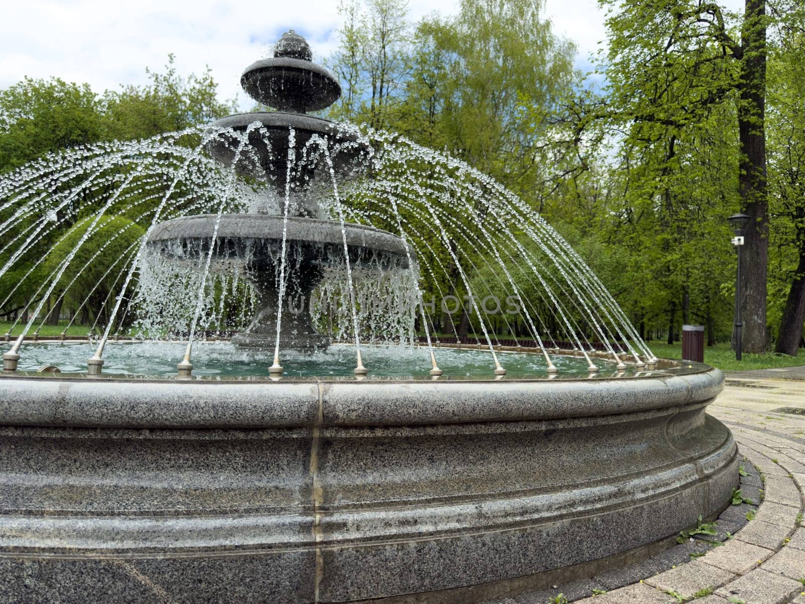 Close up of stone fountain with layered tiers and arched water jets in public park. Clear focus on textured surface and dynamic water flow design