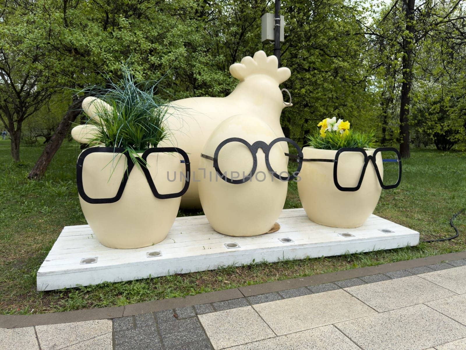 Decorative sculpture of hen and three egg shaped flower pots with black glasses on wooden platform in city park. Whimsical urban art installation with humorous design