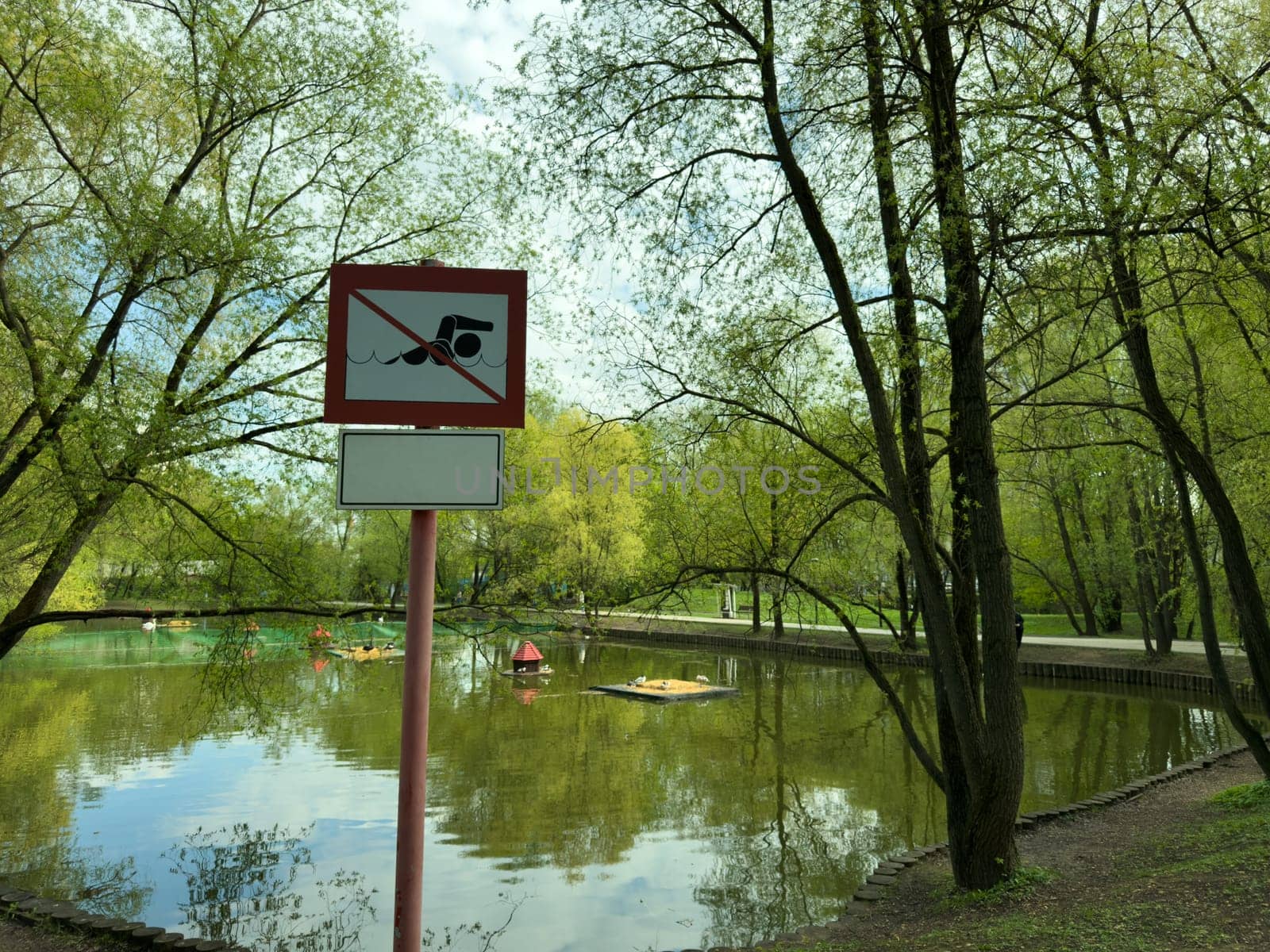 Warning sign by scenic pond in public park prohibiting swimming. The calm water reflects the surrounding trees, and area features pedal boats and small island for ducks