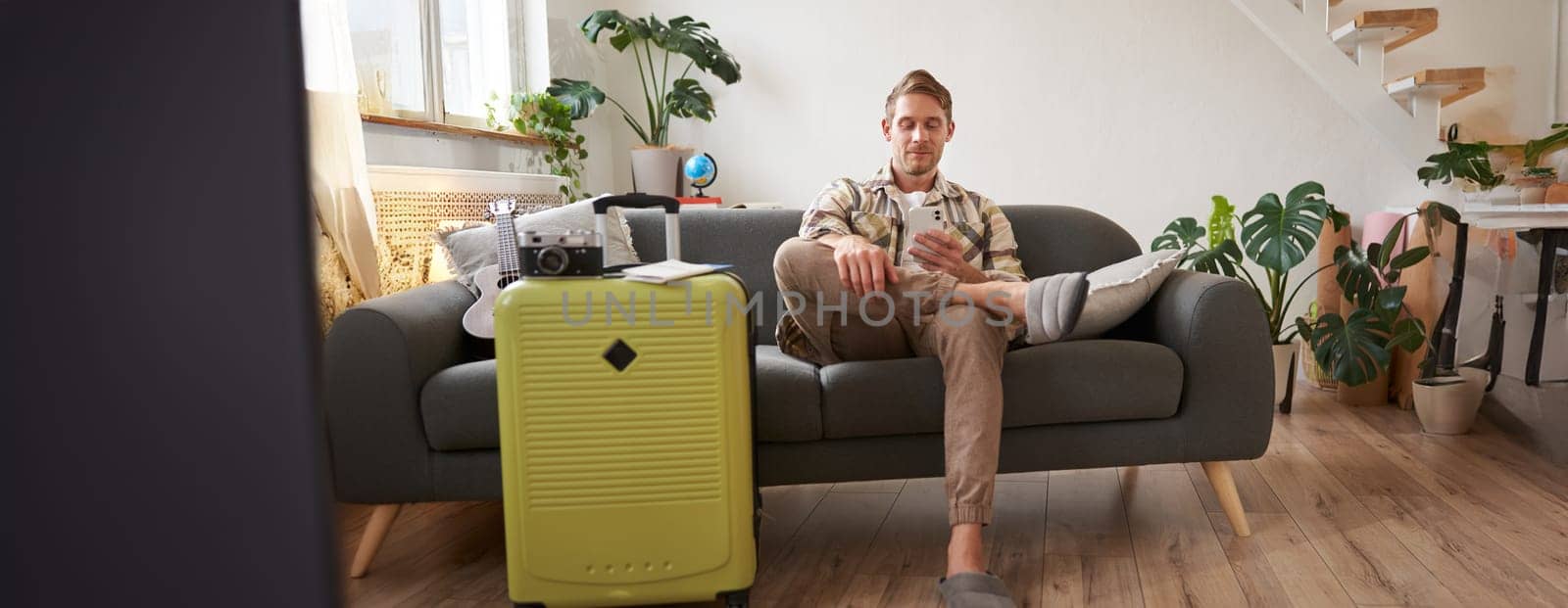 Portrait of man sits with mobile phone in living room, packed suitcase, chats on smartphone app, looks for holiday destinations, goes on vacation.