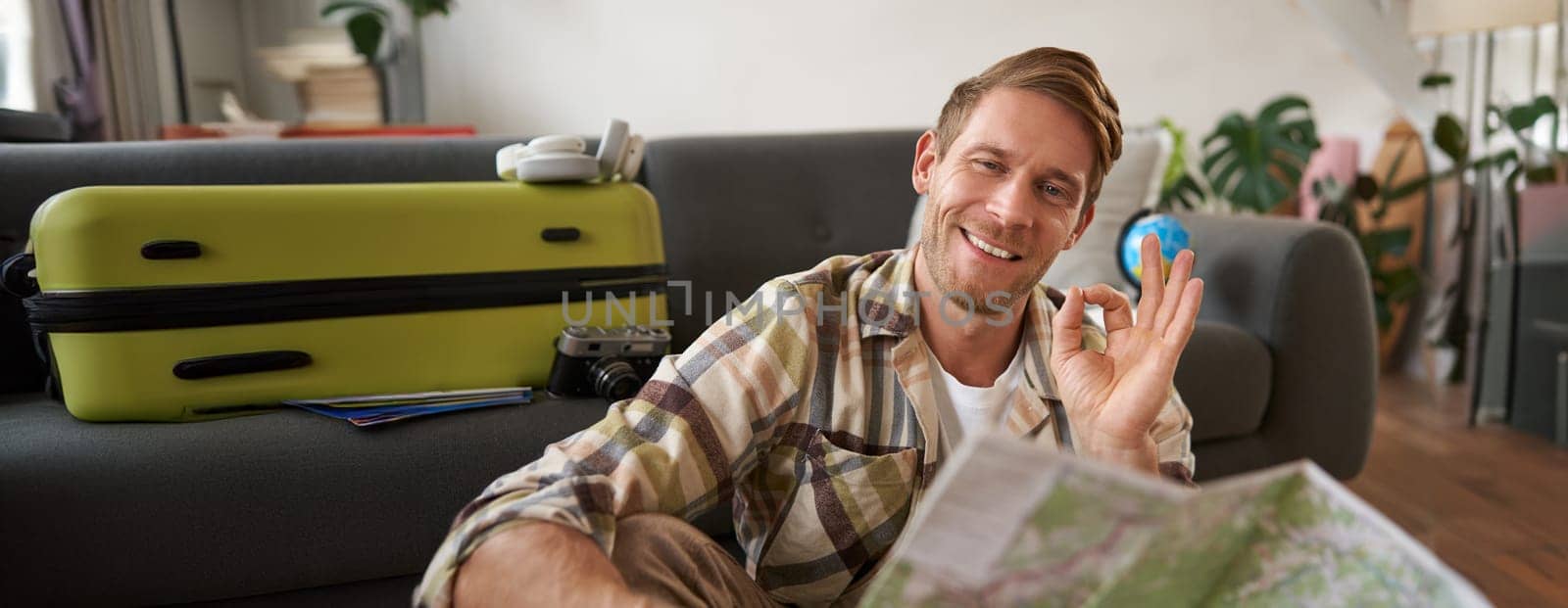 Smiling handsome tourist, man with road travel map, sits on floor near suitcase, shows okay, ok sign, recommending a tour.