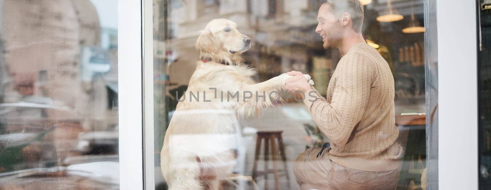 An outdoor shot of a man sitting with his dog in a pet-friendly cafe near window, golden retriever gives paw to visitor.