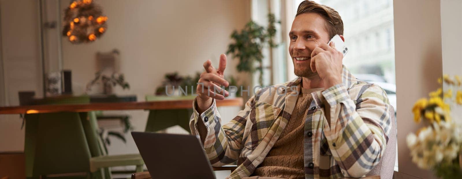 Handsome businessman in coffee shop, sitting in cafe with laptop and calling someone, Man talking on the phone with client, making enquiry about project, talking to coworker while working remotely.