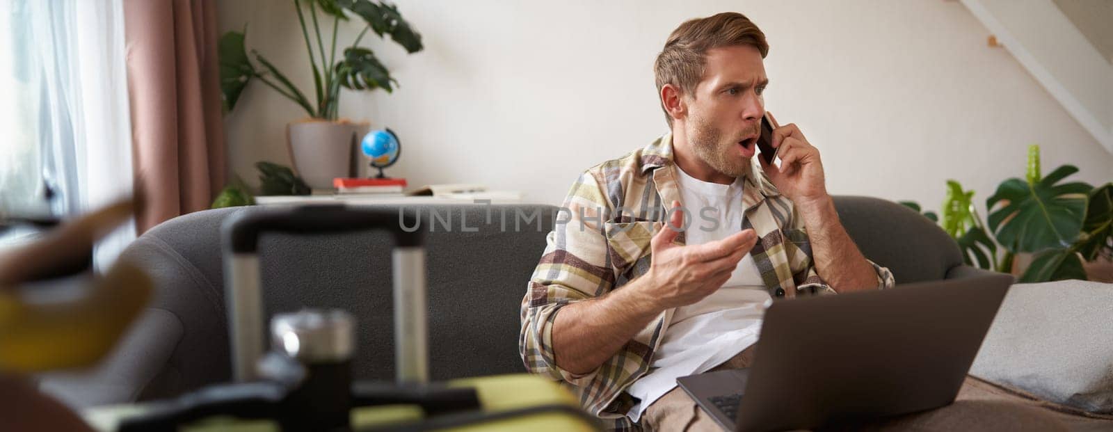 Portrait of angry man arguing over the phone, has laptop opened on his laptop, sits near suitcase, complaining at travel agency. Copy space