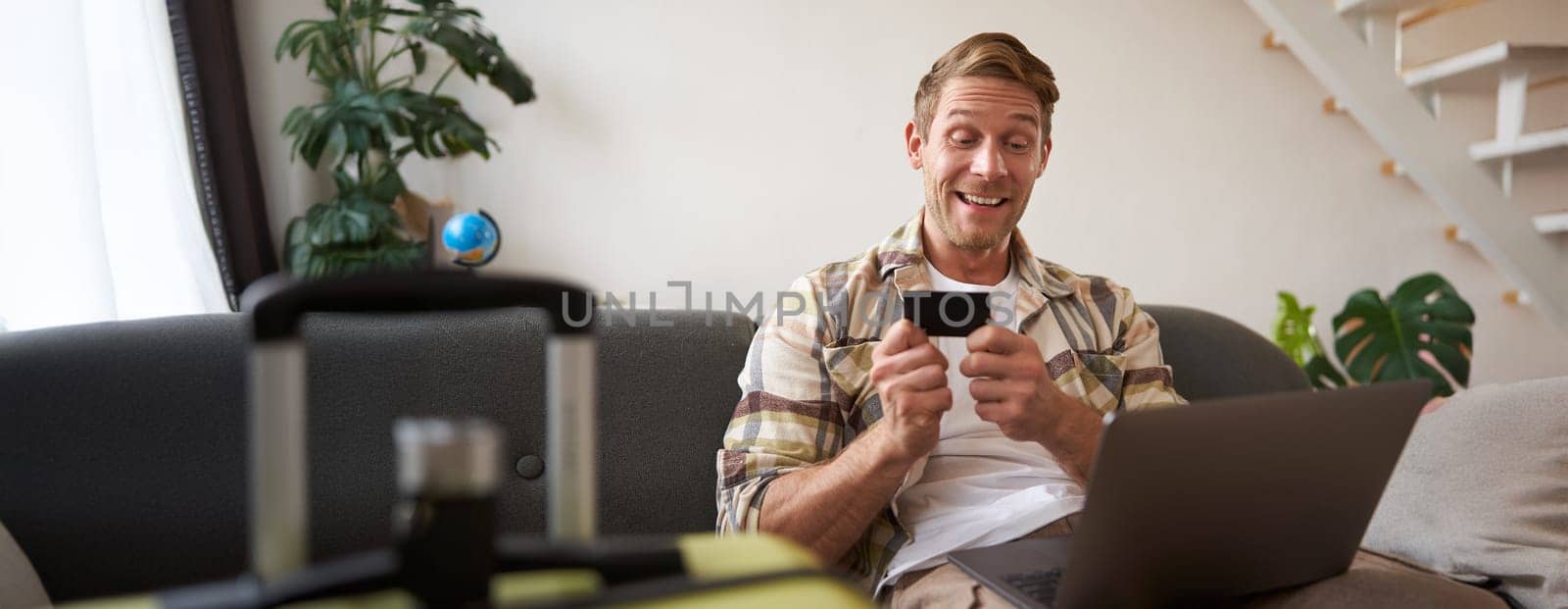 Portrait of cheerful man, going on holiday, plans a travel journey abroad, has laptop opened, sits in living room with suitcase, books a hotel.