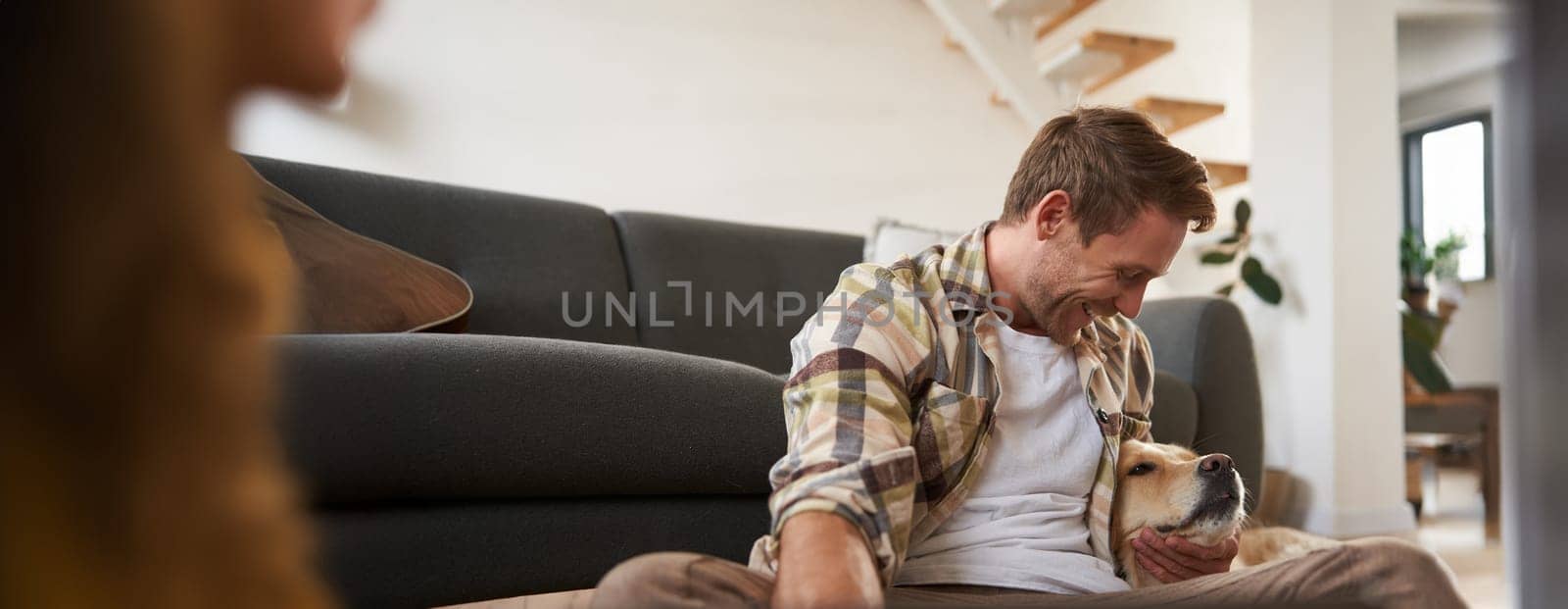 Happy young smiling guy in living room, sitting on floor, hugging his pet, playing with dog. People and animals concept