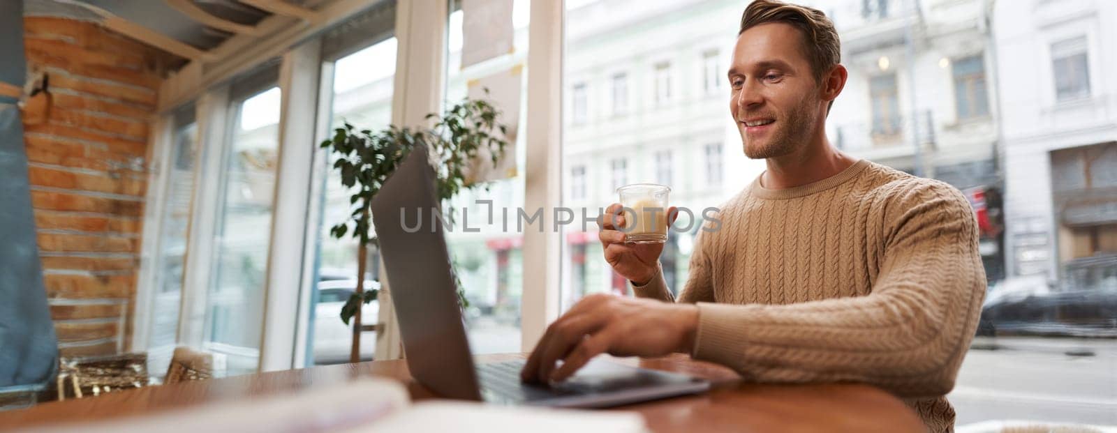 Portrait of young male businessman in cafe, drinking coffee and using his laptop, working from co-working space, sitting near the window and looking at computer screen.