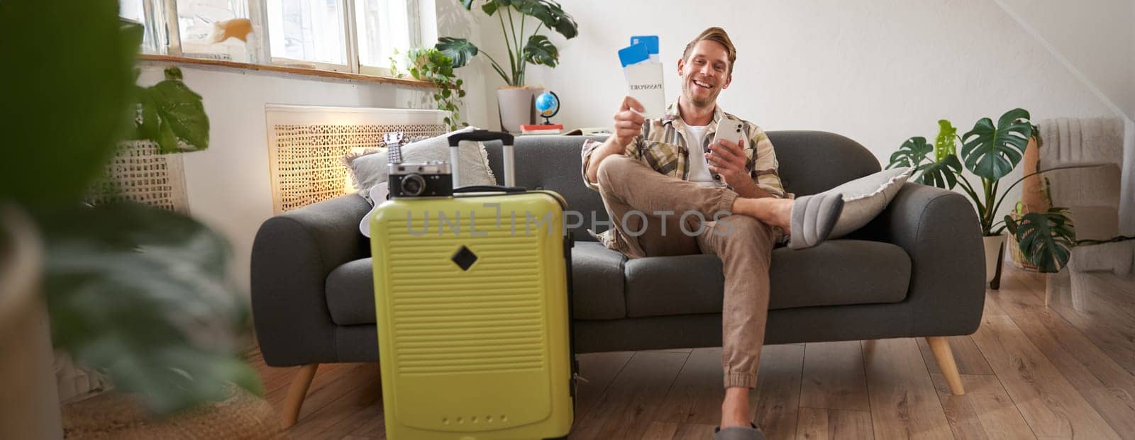 Portrait of young tourist, man on sofa with suitcase, preparing for holiday, sitting with smartphone, booking hotel and plane tickets online on mobile phone app. Tourism and travelling concept