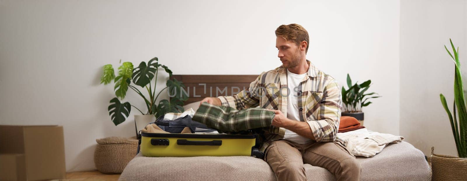 Portrait of young man packing his luggage for a trip, going on holiday, sitting on bed and putting clothes in suitcase, preparing for his flight.