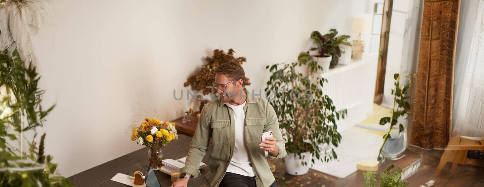 Portrait of young happy man, businessman in office, leaning on table, holding mobile phone, looking at laptop, checking project chart on screen, making a call.
