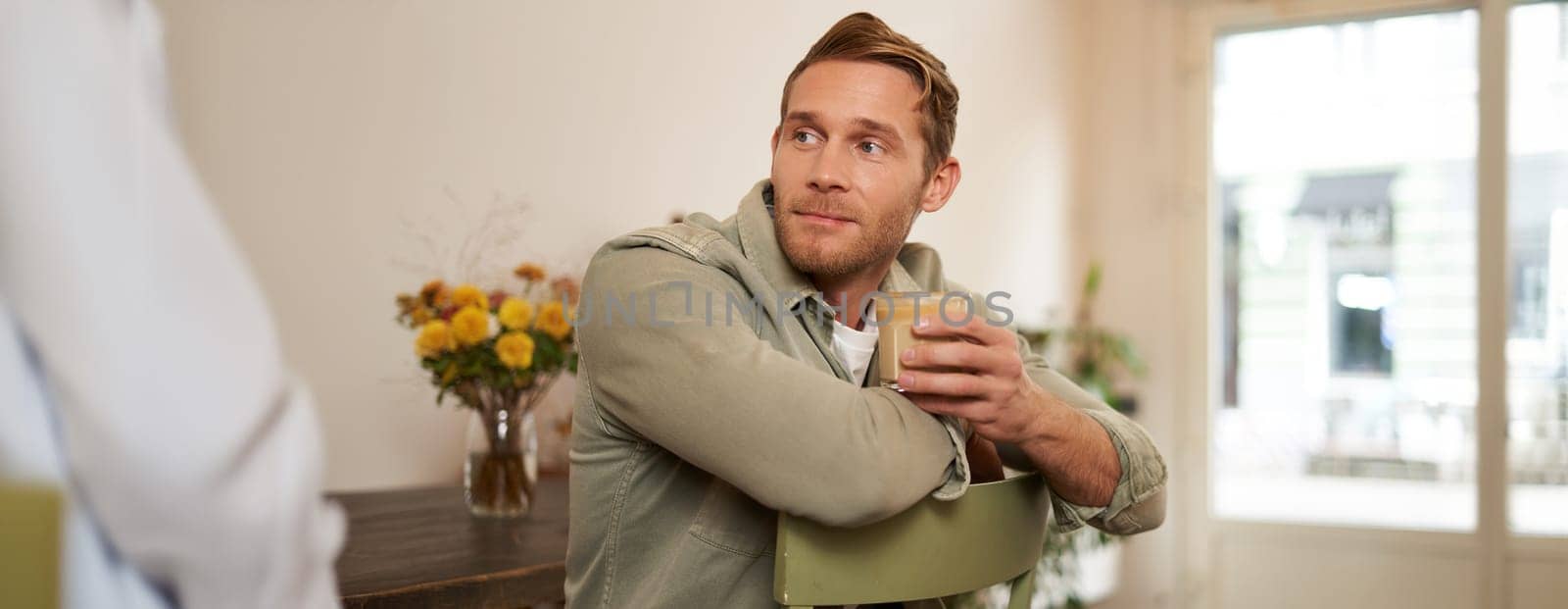 Portrait of good-looking young man with cup of coffee, sitting on chair in cafe, smiling and relaxing with his cappuccino drink.