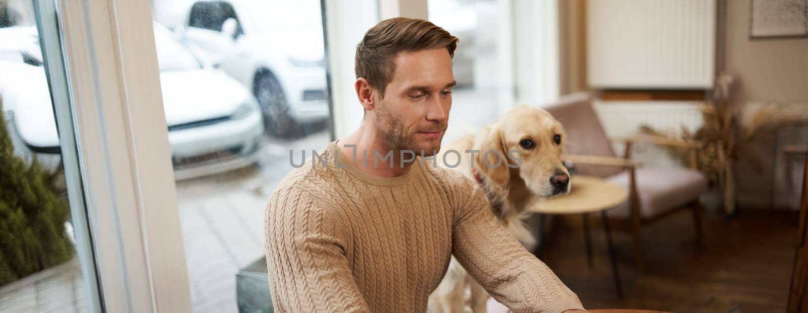 Handsome young man, digital nomad sitting with his dog in a pet-friendly cafe, working on laptop while a golden retriever watches him.