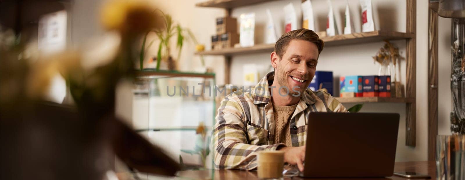 Portrait of handsome young man using laptop in cafe, sitting with computer in creative public space, typing on keyboard with pleased happy smile.
