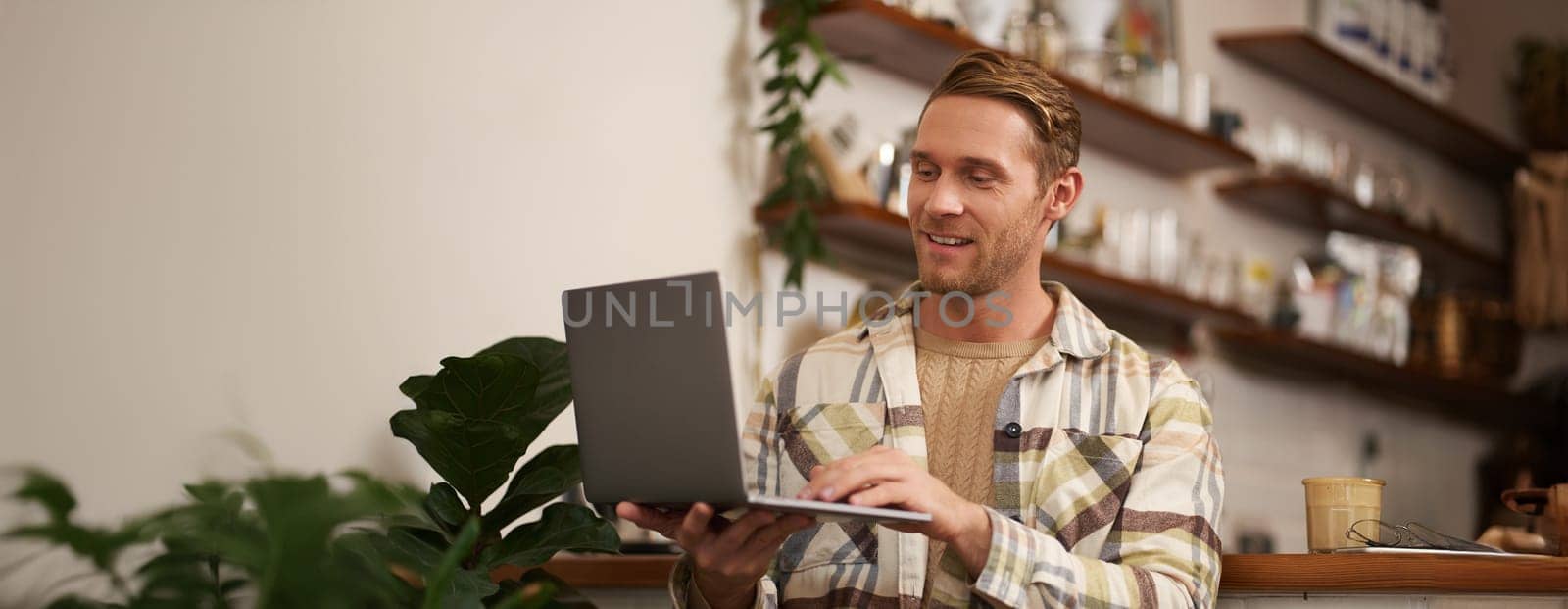 Portrait of young man working online, freelancer sitting in cafe, doing his project on laptop, drinking coffee, smiling while looking at screen.