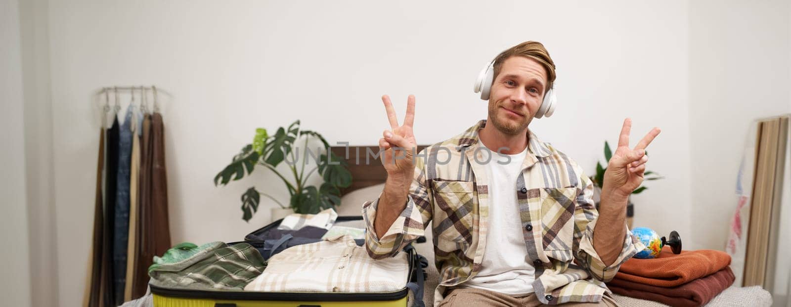 Portrait of cheerful handsome man, tourist going on vacation, dancing and listening to music in headphones while packing suitcase with clothes for holiday trip abroad.