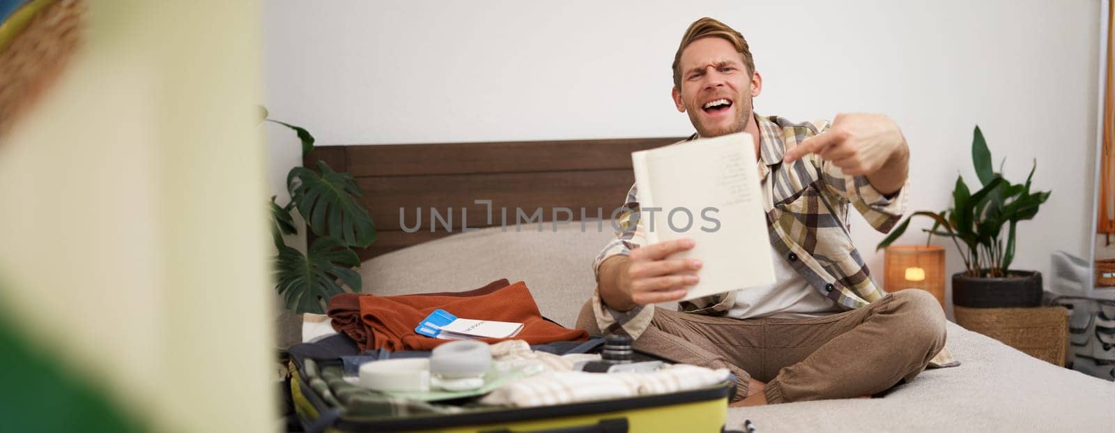 Travelling and tourism. Young man sits on bed, shows his list of items, demonstrates notebook, packs clothes, prepares to go on holiday, goes on vacation, assembles luggage.