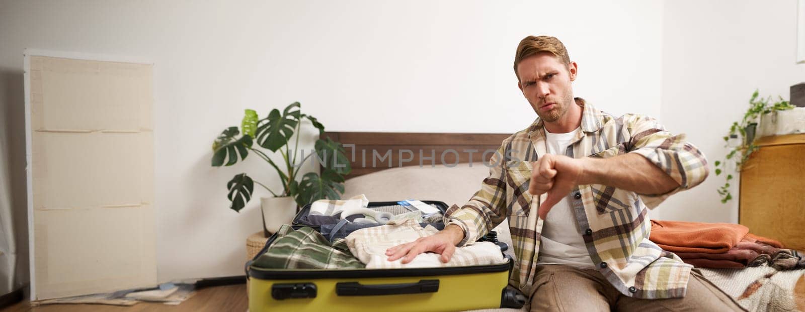 Image of young disappointed man, showing thumbs down, puts hand on his clothes inside luggage, dislike, disapprove something awful, upset with his holiday. Lifestyle concept
