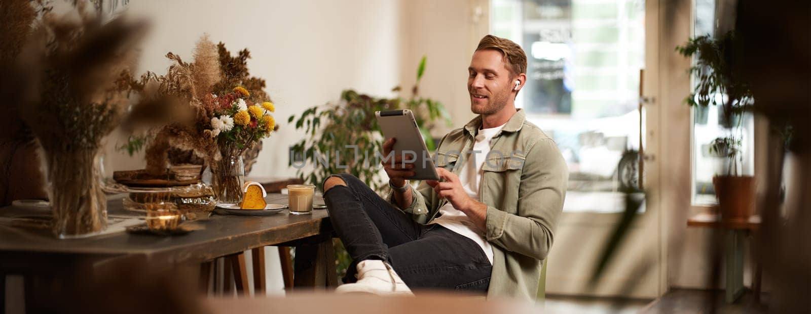 Portrait of young man sitting in cafe, spending time alone in coffee shop, reading on digital tablet, watching video, listening music in wireless headphones. Lifestyle and people concept.