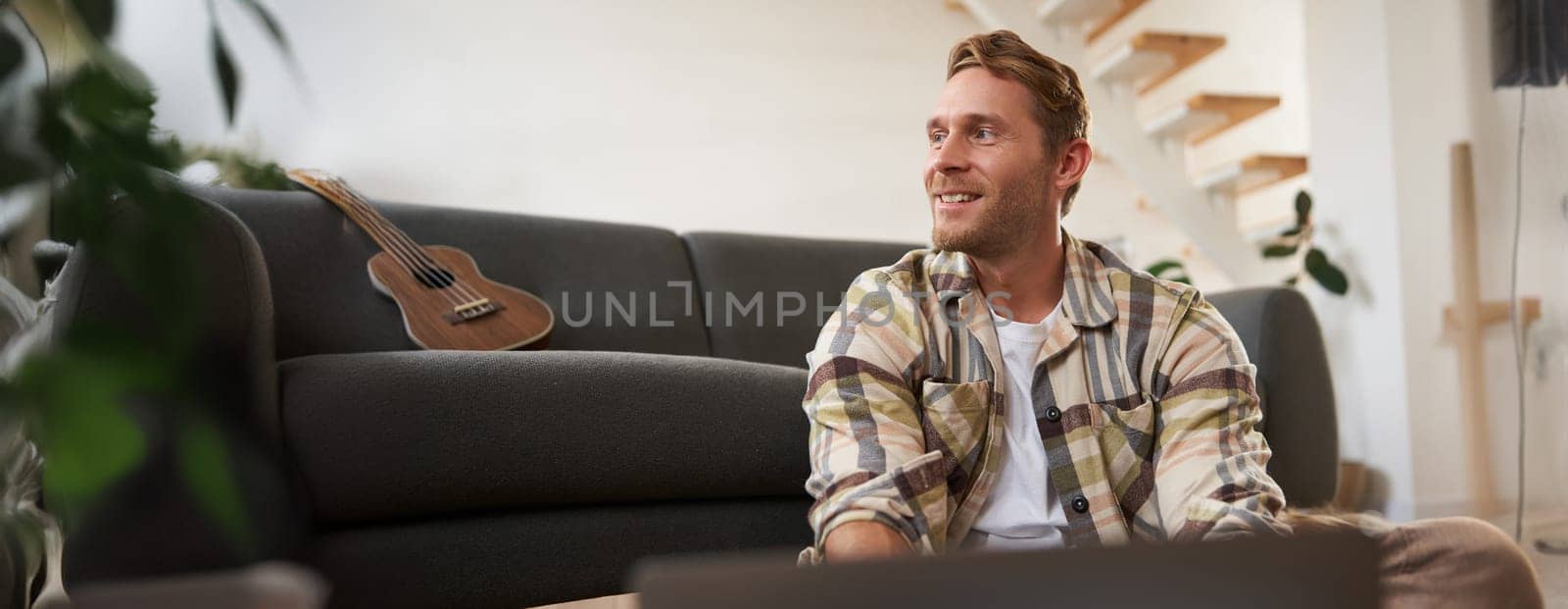 Close up portrait of young man using laptop, looking at computer, sitting on floor with his gadget, browsing websites.