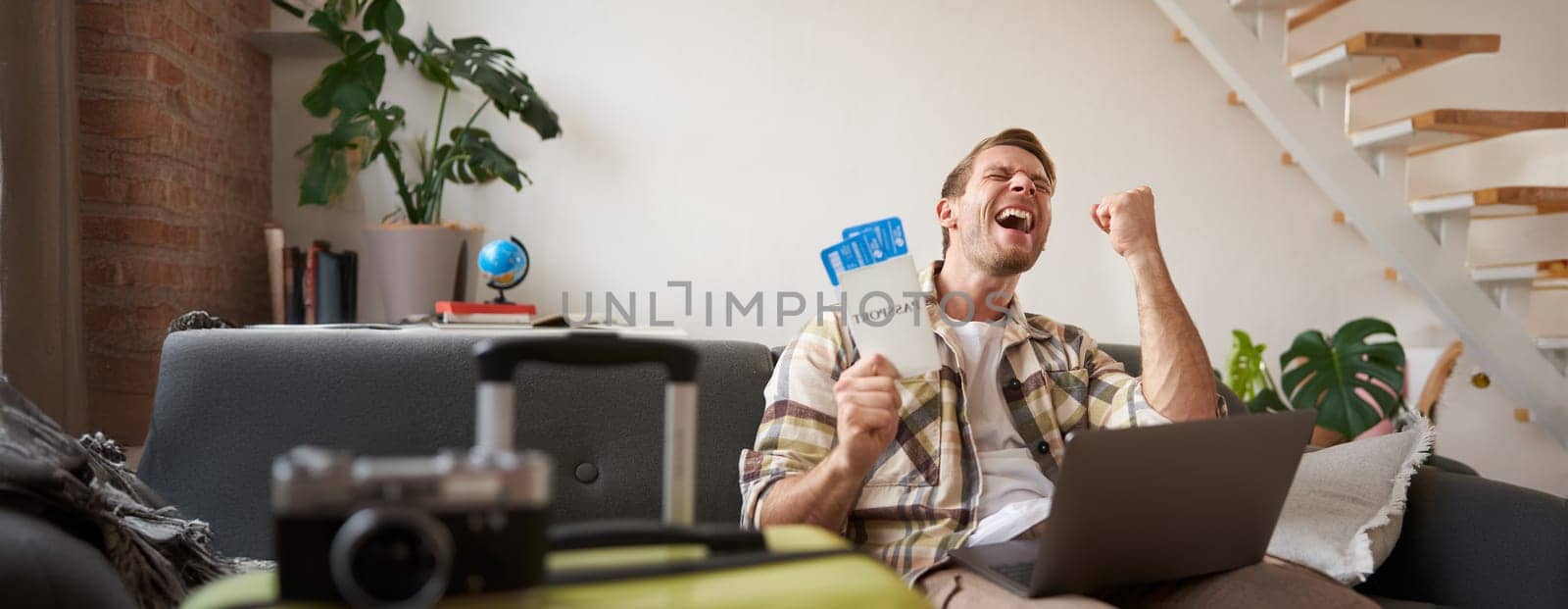 Enthusiastic young man, tourist looking excited, saying yes, holding two tickets, sitting with laptop and screaming from happiness, has suitcase and camera prepared for holiday.