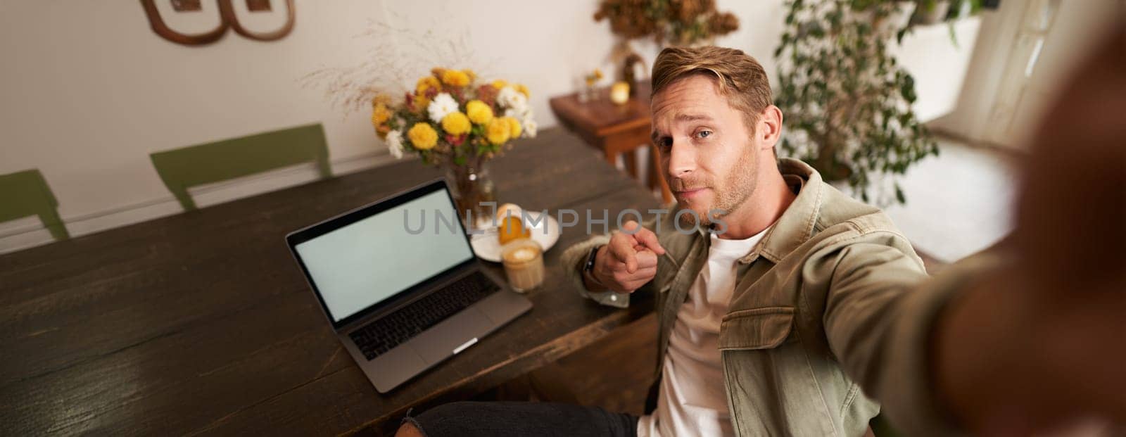 Handsome and confident young man taking selfie on mobile phone, pointing finger at camera, sitting in cafe with laptop, working from coffee shop, looking sassy.
