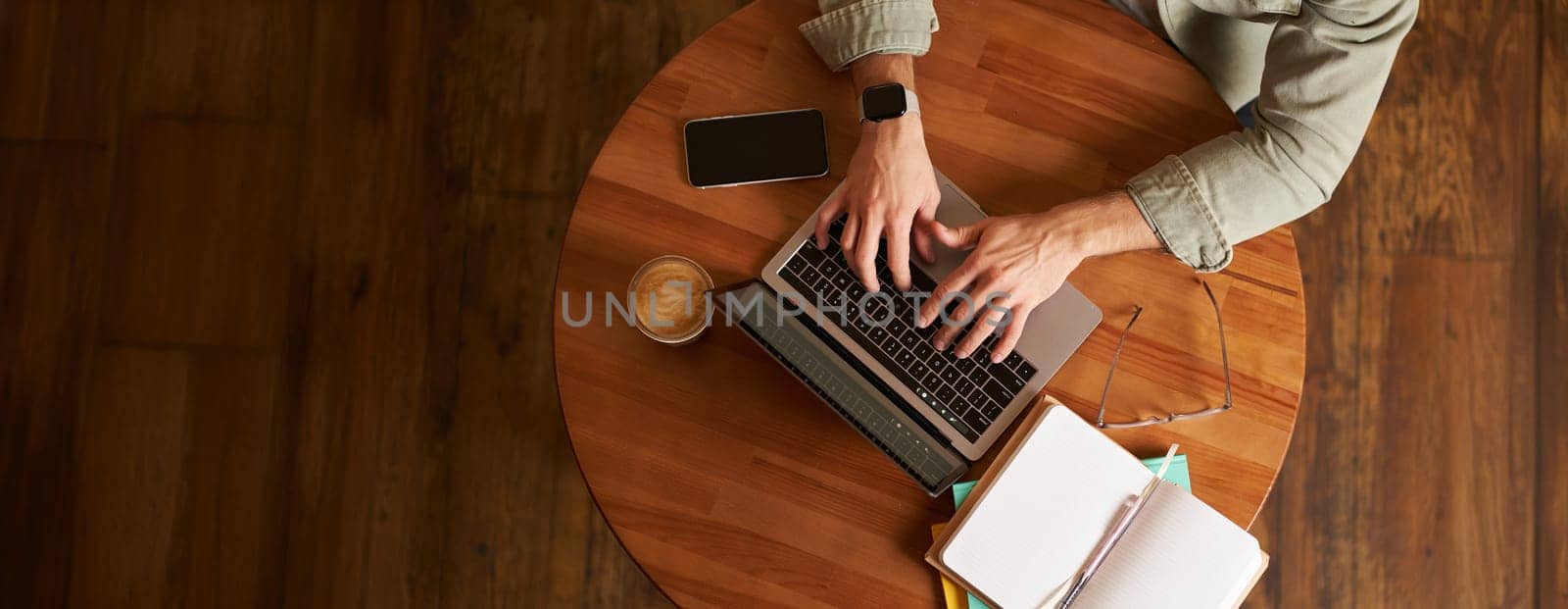 Cropped top view of male hands, man typing on laptop keyboard, working in cafe, sitting at round table in coffee shop, has phone, notebook and cup.