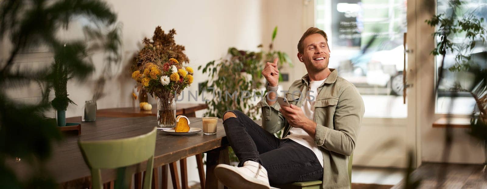 Portrait of young handsome man in casual clothes, sits in cafe and enjoys favourite song, listens to music in wireless headphones, vibing to the relaxing tunes in earphones, holding smartphone.