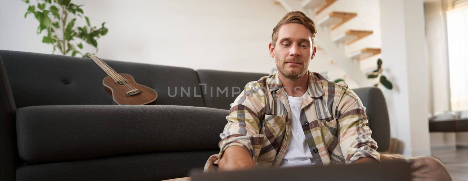 Close up portrait of young man using laptop, looking at computer, sitting on floor with his gadget, browsing websites.