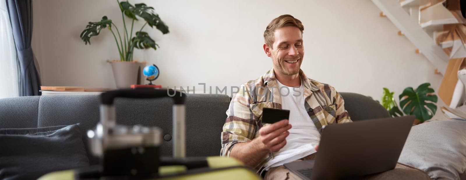Happy man booking holiday online on laptop, paying for vacation on travel agency website, buying flight tickets in internet, sitting in living room with suitcase.