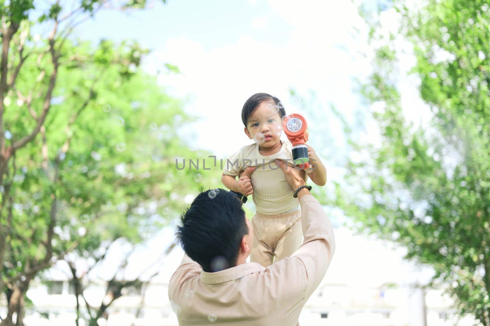 Happy child holding a bubble toy while being lifted by dad in the park.