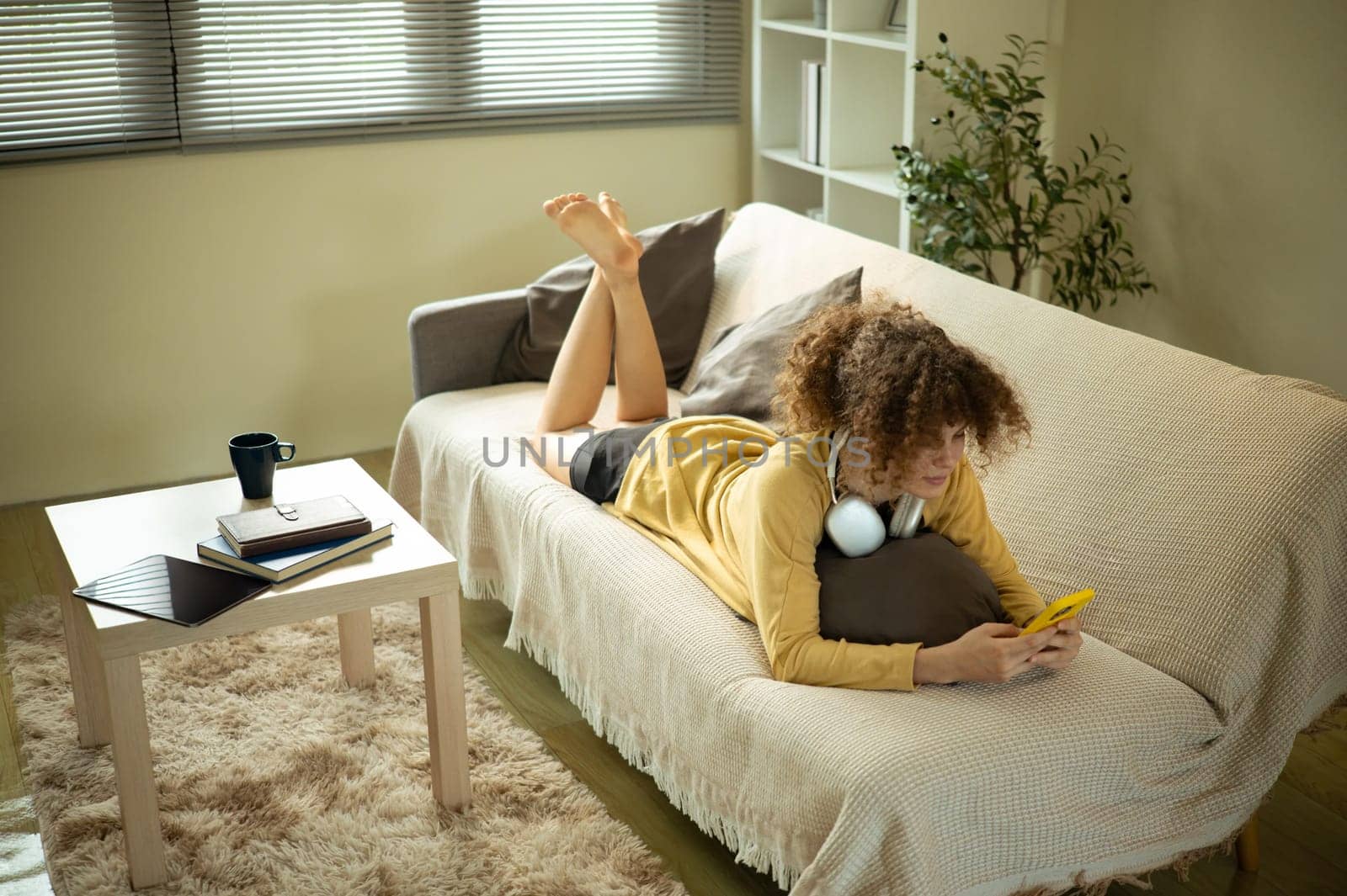 Young woman lying on sofa and using smartphone in a cozy living room.