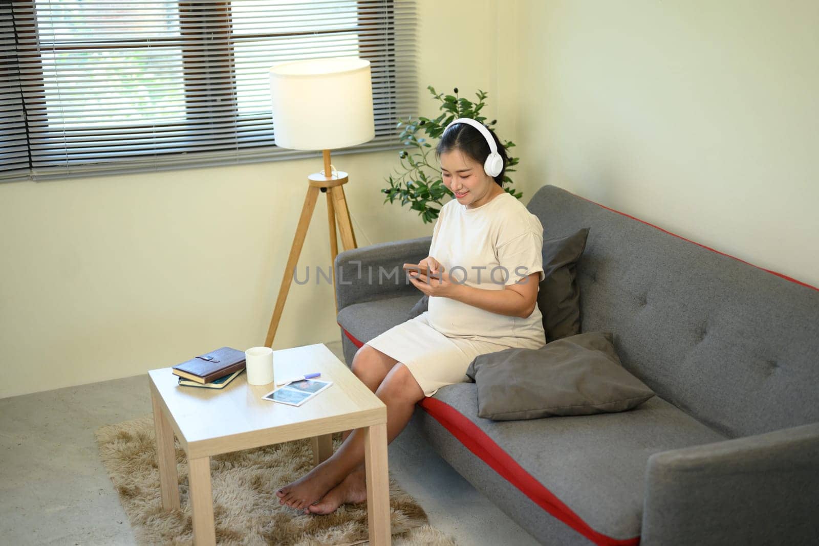 Pregnant woman relaxing on sofa, listening to music with headphones and using a smartphone.