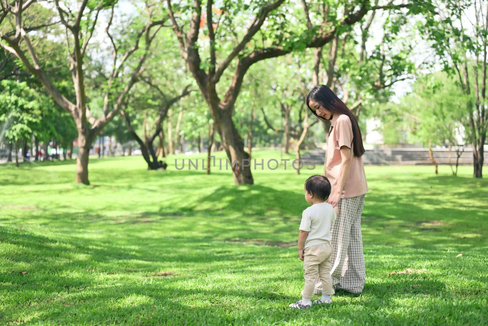 Mother and toddler holding hands walking through a peaceful green park on a sunny day.